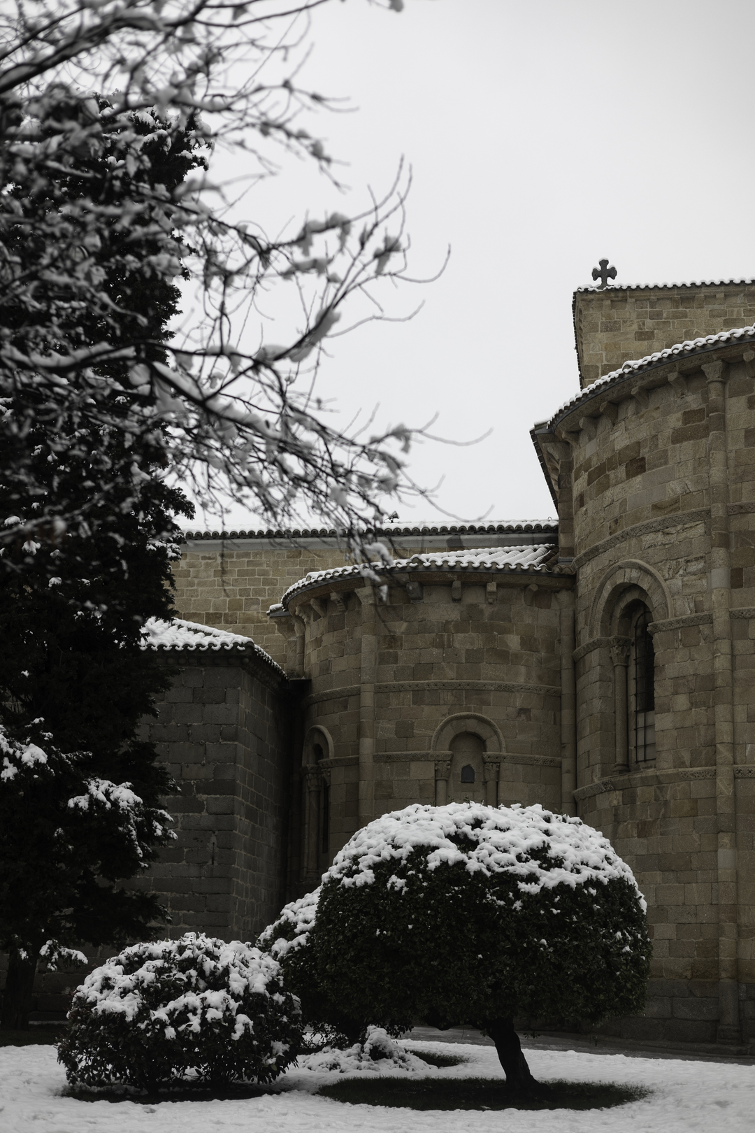 Heavy snow blankets the garden bushes outside the rear Romanesque exterior of San Pedro Church in Avila, Spain, highlighting the ancient stone architecture on a freezing winter day. (c) pmartinasi