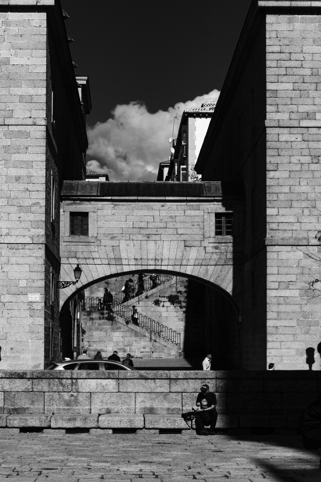  Capturing the architectural charm of San Lorenzo de El Escorial, featuring a stone archway that connects two imposing stone buildings. Above the arch sits a bridge-like structure with a small window, while below, people ascend and descend a staircase that leads through the archway. The interplay of historic textures, light, and shadow contrasts beautifully with modern buildings visible in the background, highlighting Spain's cultural layers and timeless urban design. (c) pmartinasi