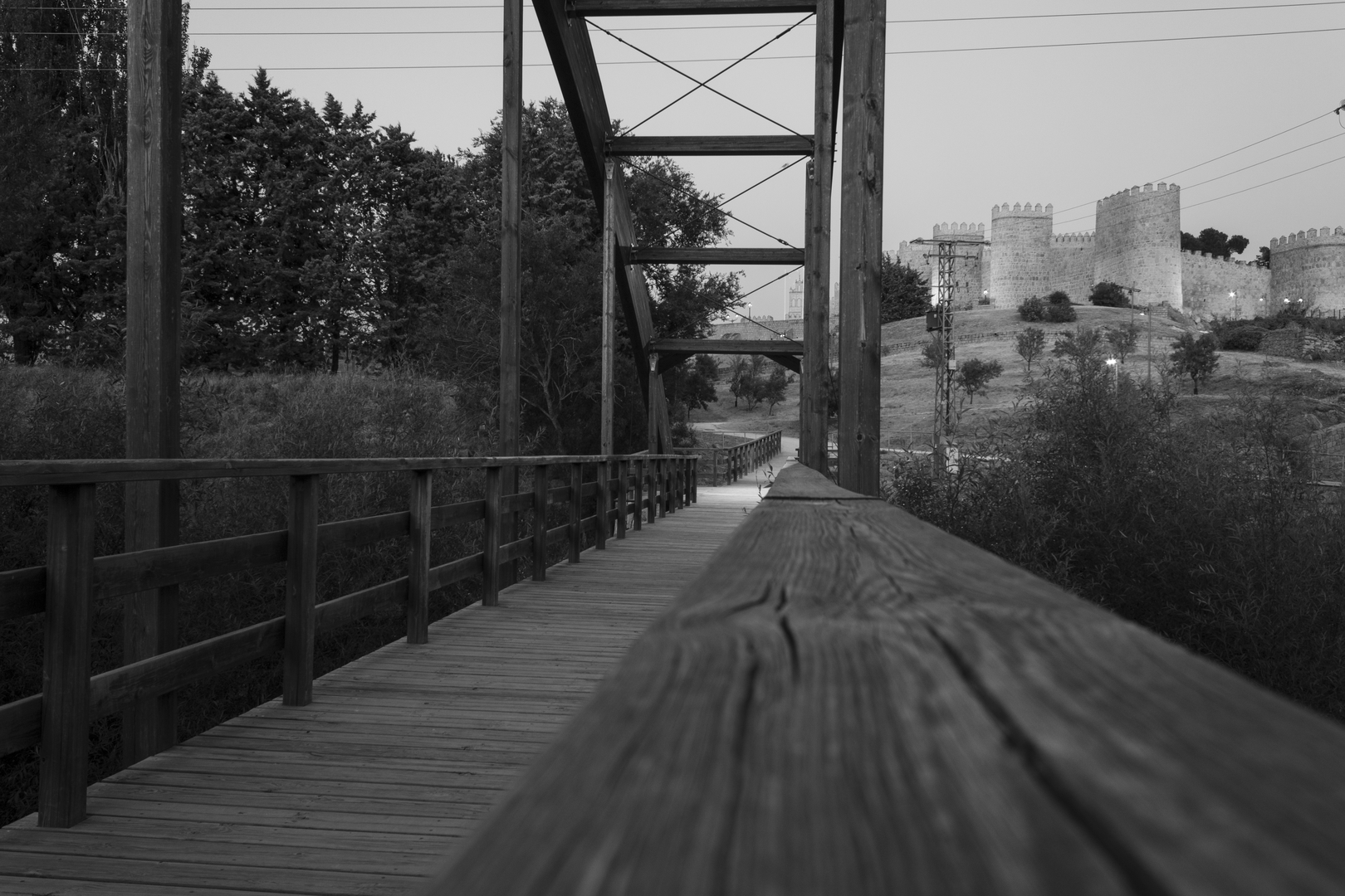  Striking black and white photograph captures the approach to Ávila's iconic medieval stone wall through a rustic wooden bridge with railings and overhead metal cross-bracing. The scene blends natural surroundings—trees and bushes—with historical architecture featuring rounded towers and fortification elements. The foreground emphasis on the bridge's railing enhances depth and perspective, creating a dramatic visual transition between nature and centuries-old Spanish heritage. (c) pmartinasi
