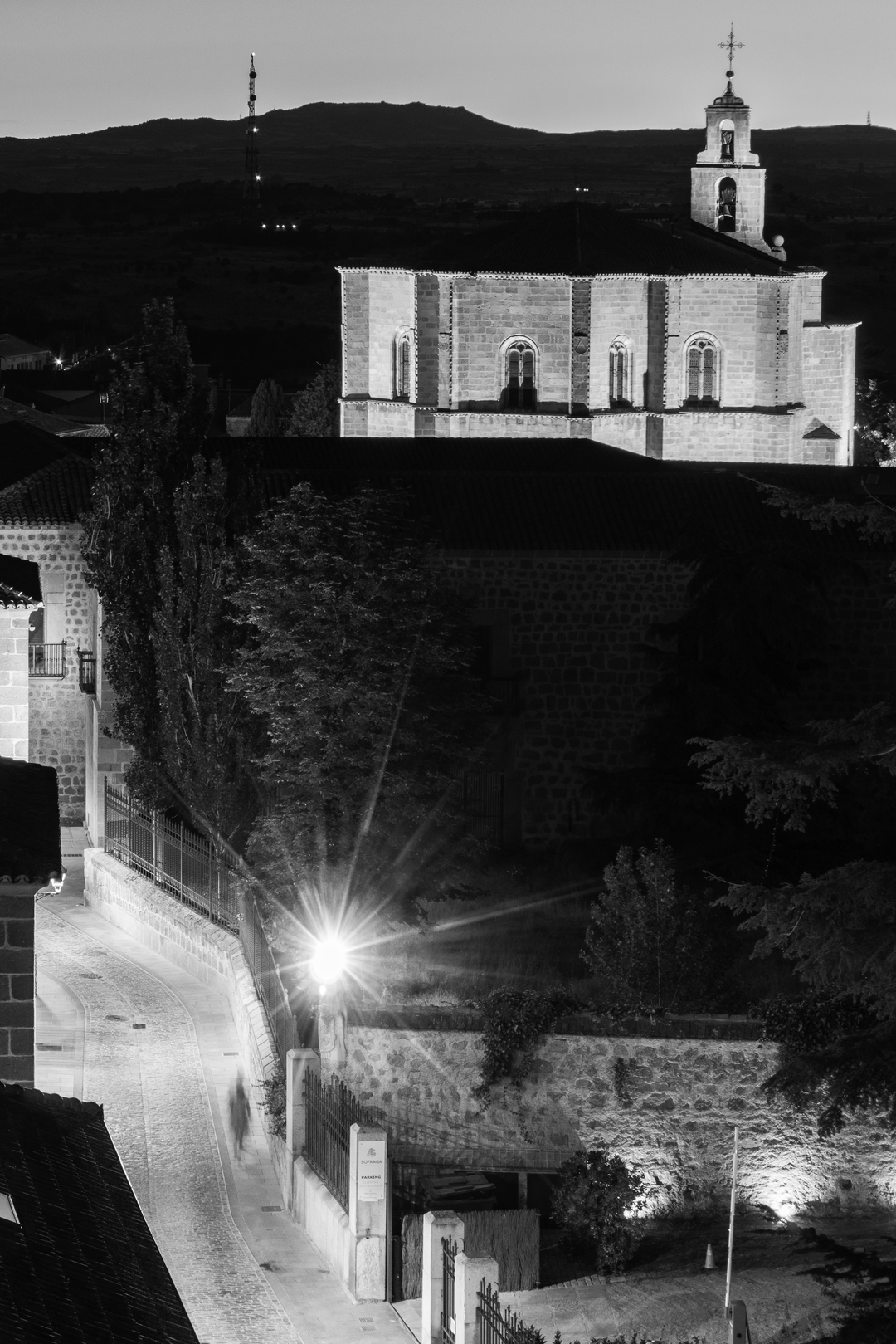  Serene majesty of the church in Mosen Rubí, Ávila, Spain. Artificial lighting reveals architectural details such as arched windows and a cross-topped bell tower, standing quietly against the softly lit hills and communication tower in the distance. Framed by trees and rooftops in the foreground, the image emphasizes the contrast between historical spirituality and modern presence under a dusky sky. (c) pmartinasi