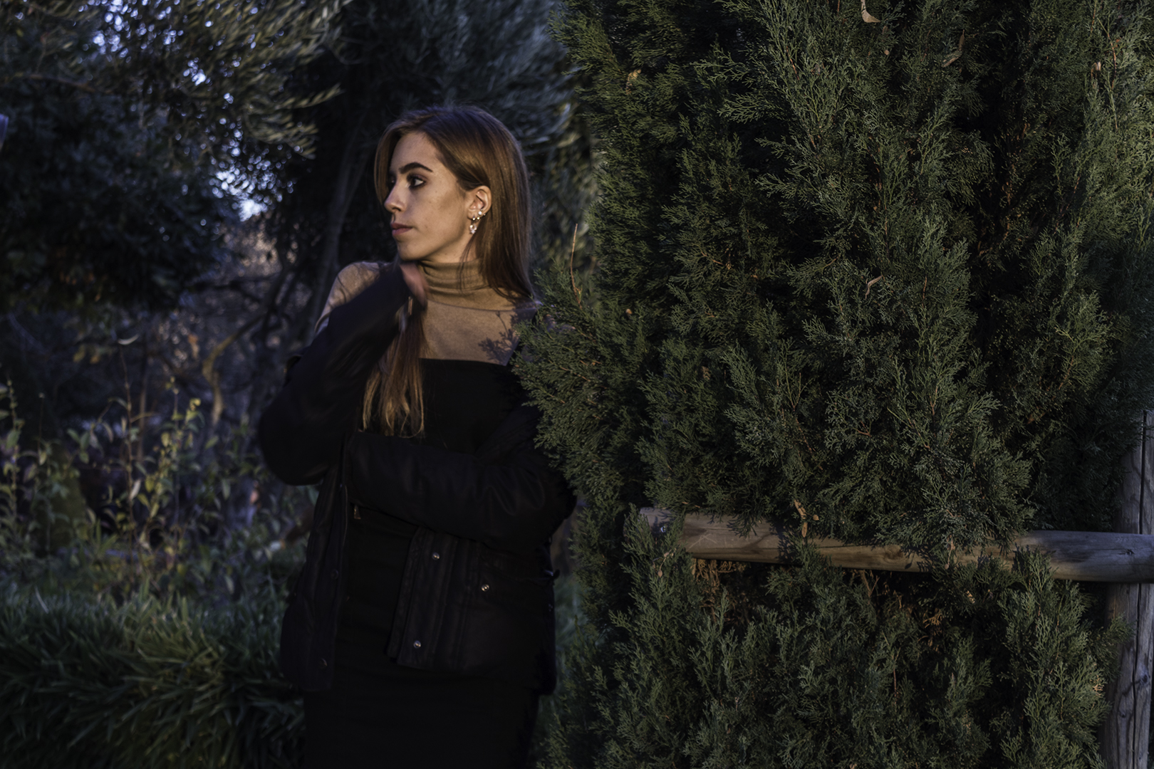 Woman in Black Jacket and Dress Standing Near Green Bush in Outdoor Setting. (c) pmartinasi
