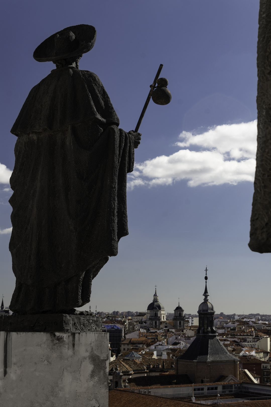  A bronze statue of Saint James (Santiago) stands atop the Cathedral of La Almudena in Madrid, Spain, facing the skyline and baroque domes of the historic city under a bright blue sky. (c) pmartinasi