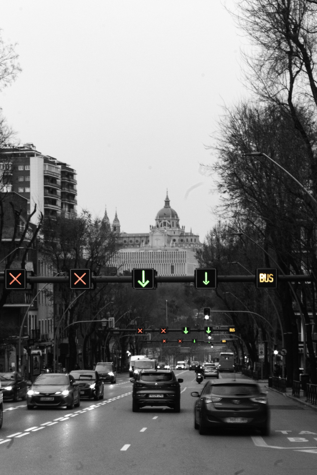 Street-level view of traffic moving through a wide avenue in Madrid, with illuminated traffic signals leading the eye towards the Almudena Cathedral in the distance, captured outdoors in a European city setting. (c) pmartinasi