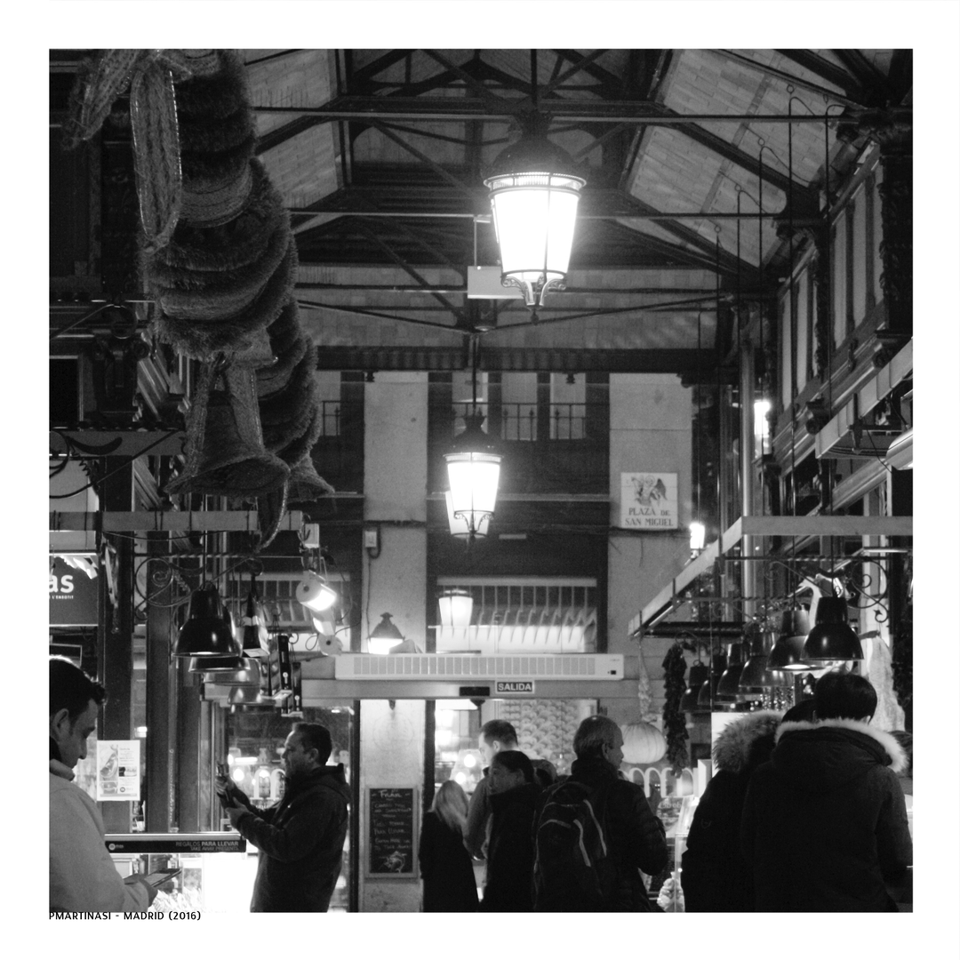  Black and white photograph of the bustling interior of Madrid's Mercado de San Miguel, highlighting the elegant ironwork, hanging cured meats, warm lighting, and lively ambience of this iconic food market. (c) pmartinasi
