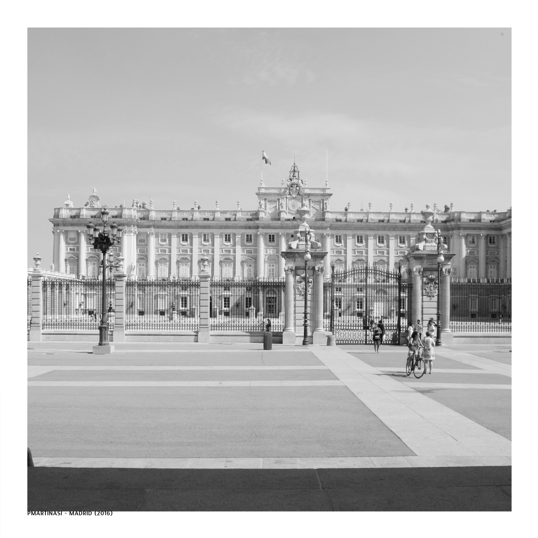  A timeless black and white photograph of the Royal Palace of Madrid (Palacio Real), capturing the grandeur of the architecture under a clear sky with pedestrians and cyclists animating the forecourt. (c) pmartinasi