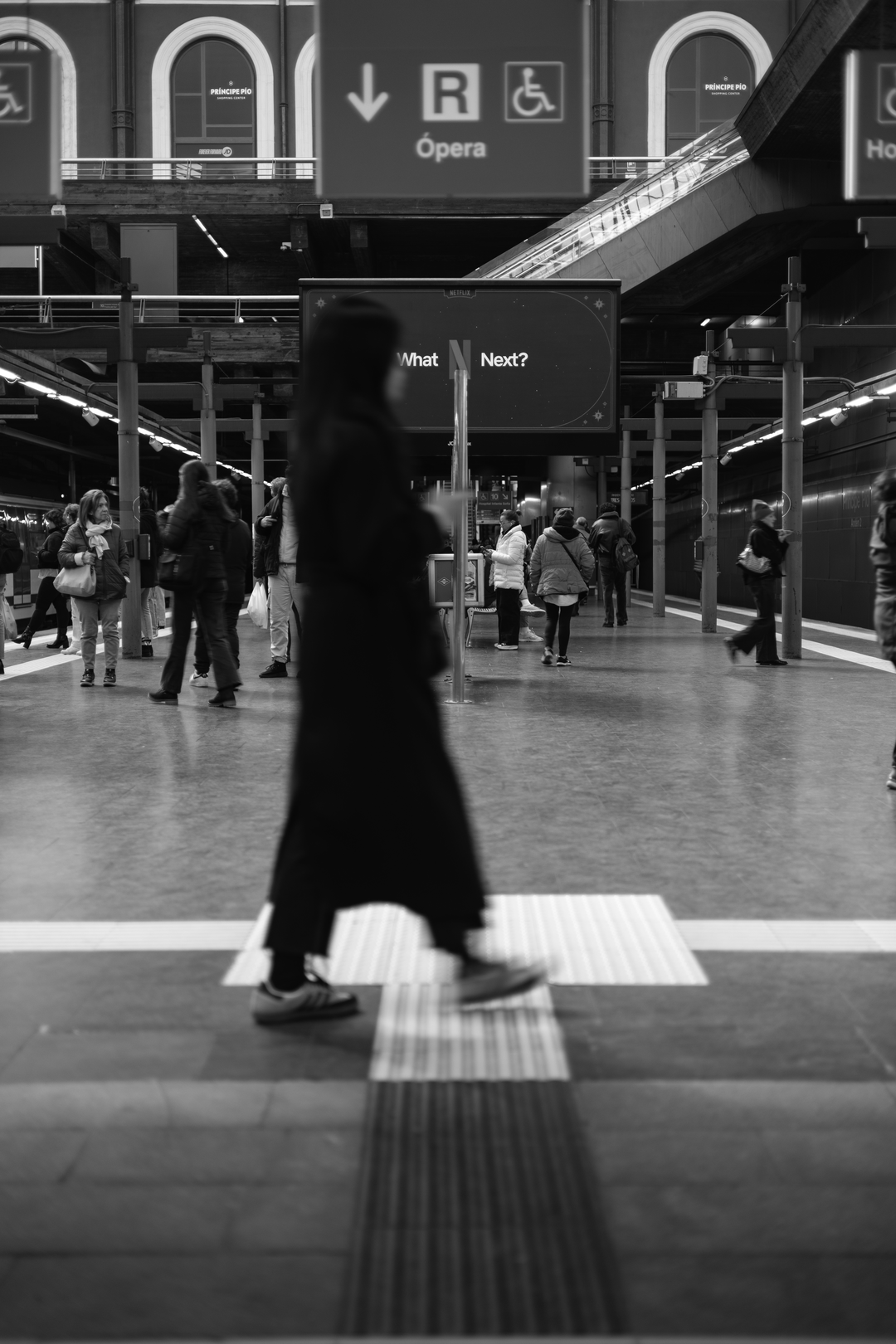 Candid black and white photograph captures a woman in motion blur walking across the platform at the busy Principe Pio Metro Station in Madrid, Spain, Europe. (c) pmartinasi