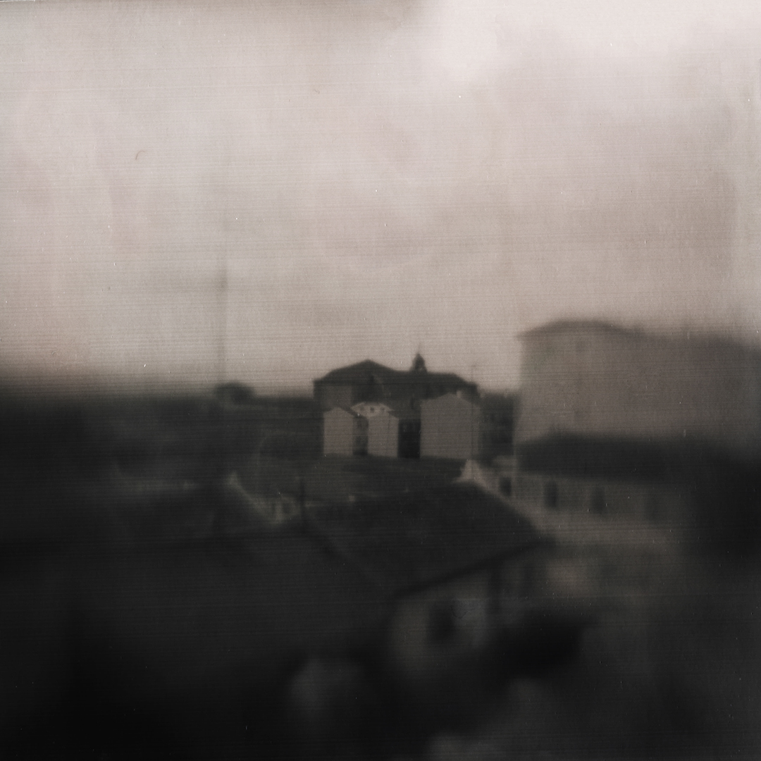 A dreamy, abstract photograph featuring a blurred view of a townscape with muted, vintage tones. The image evokes nostalgia and serenity, capturing a timeless rural atmosphere. (c) pmartinasi