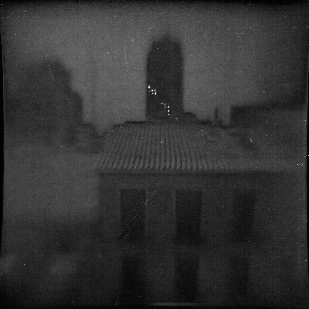Atmospheric black-and-white photograph showcasing a blurred view of the Plaza España skyline in Madrid. The moody composition features rooftops and a prominent building in the distance, evoking nostalgia and urban solitude. Camera design by @joterman. (c) pmartinasi