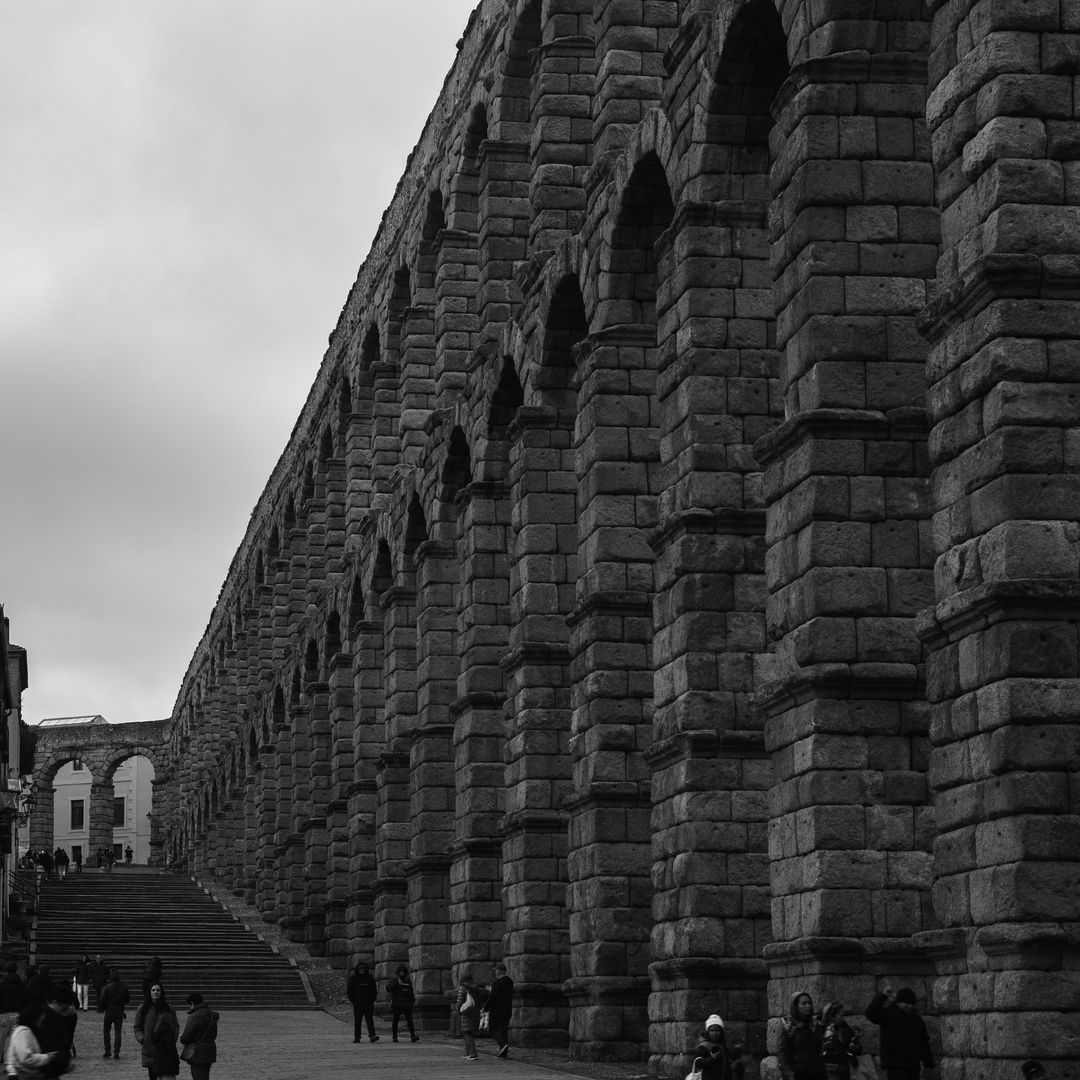 Imposing perspective view of the ancient Roman stone Aqueduct of Segovia, a historic monument in Segovia, Spain, with people walking beneath the granite arches. (c) pmartinasi