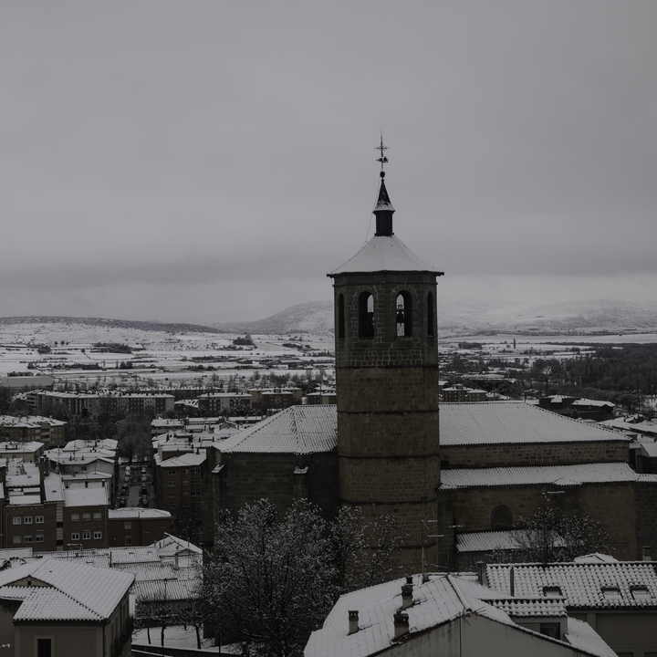 An elevated winter view overlooking the snowy Church of Santiago the Apostle and residential rooftops in southern Avila, Spain, with the snow-capped Gredos Mountains visible in the distance under an overcast sky. (c) pmartinasi