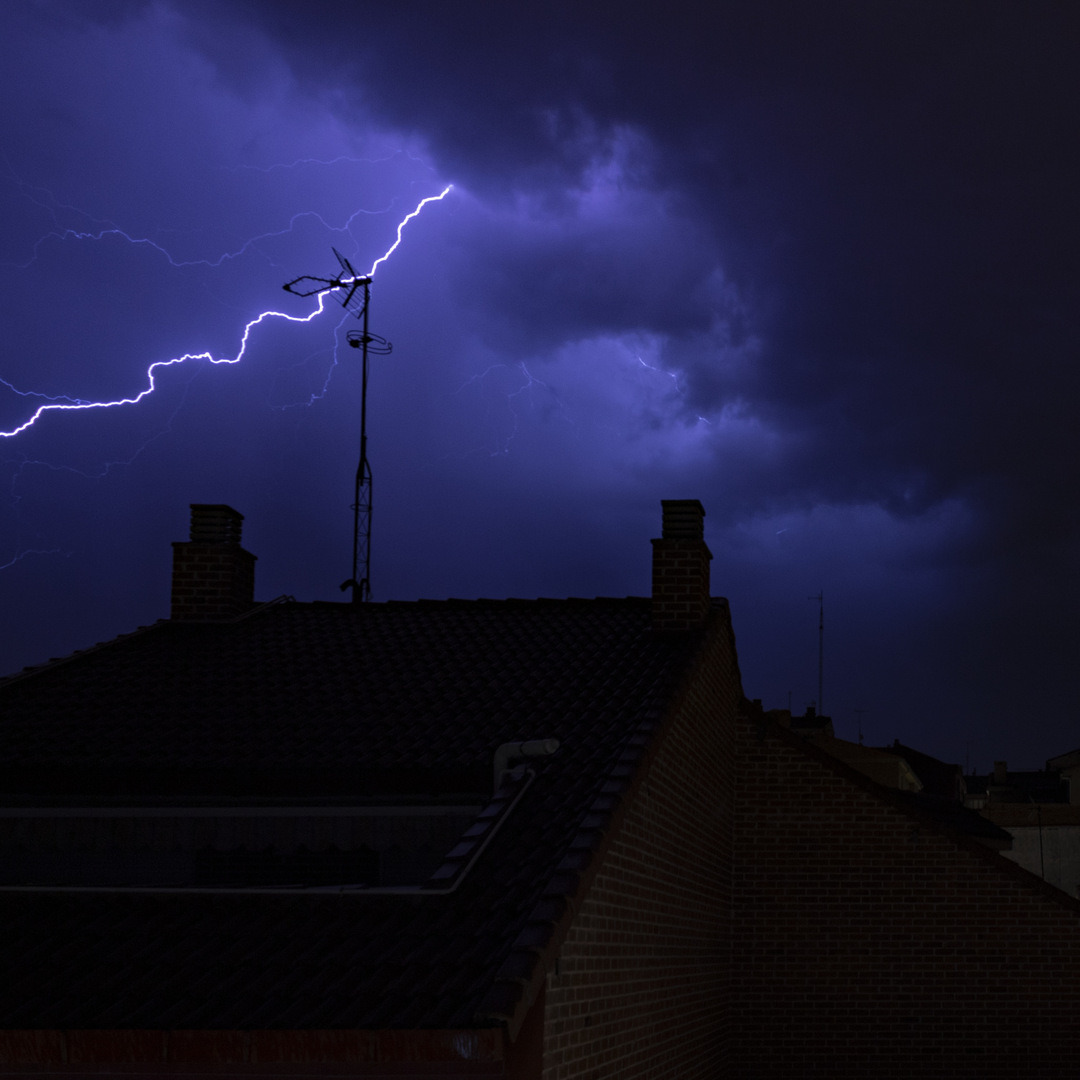  A dramatic lightning bolt slashes across a dark, cloud-covered night sky, casting eerie light over a rooftop featuring two brick chimneys and a TV antenna. The electric energy and looming clouds highlight the awe and danger of nature's power, with the antenna seemingly within reach of the strike—emphasizing structural vulnerability during thunderstorms.. (c) pmartinasi