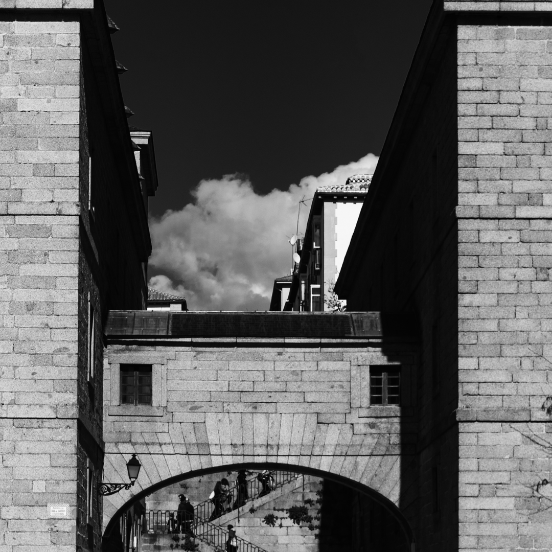  Capturing the architectural charm of San Lorenzo de El Escorial, featuring a stone archway that connects two imposing stone buildings. Above the arch sits a bridge-like structure with a small window, while below, people ascend and descend a staircase that leads through the archway. The interplay of historic textures, light, and shadow contrasts beautifully with modern buildings visible in the background, highlighting Spain's cultural layers and timeless urban design. (c) pmartinasi