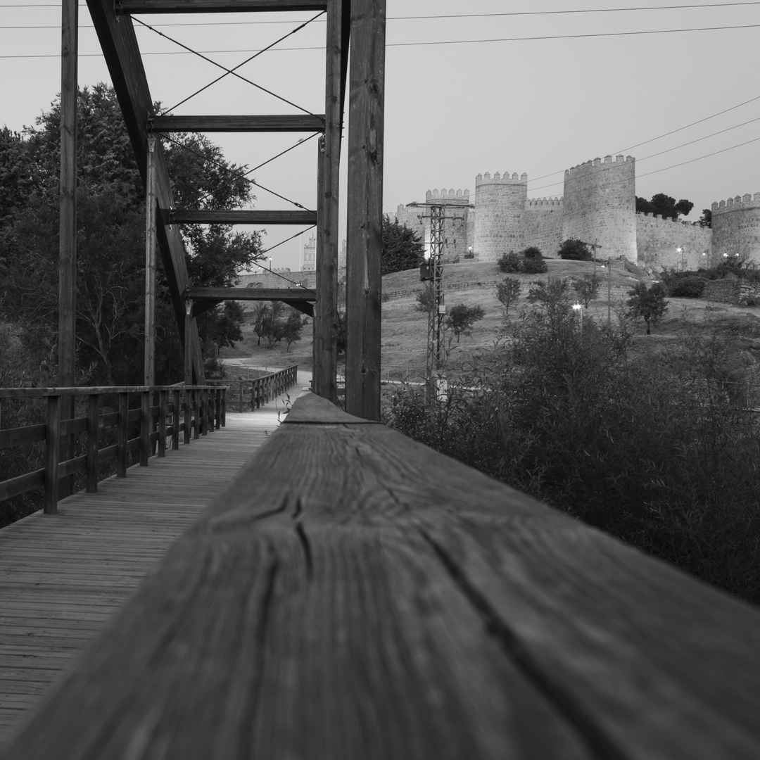  Striking black and white photograph captures the approach to Ávila's iconic medieval stone wall through a rustic wooden bridge with railings and overhead metal cross-bracing. The scene blends natural surroundings—trees and bushes—with historical architecture featuring rounded towers and fortification elements. The foreground emphasis on the bridge's railing enhances depth and perspective, creating a dramatic visual transition between nature and centuries-old Spanish heritage. (c) pmartinasi