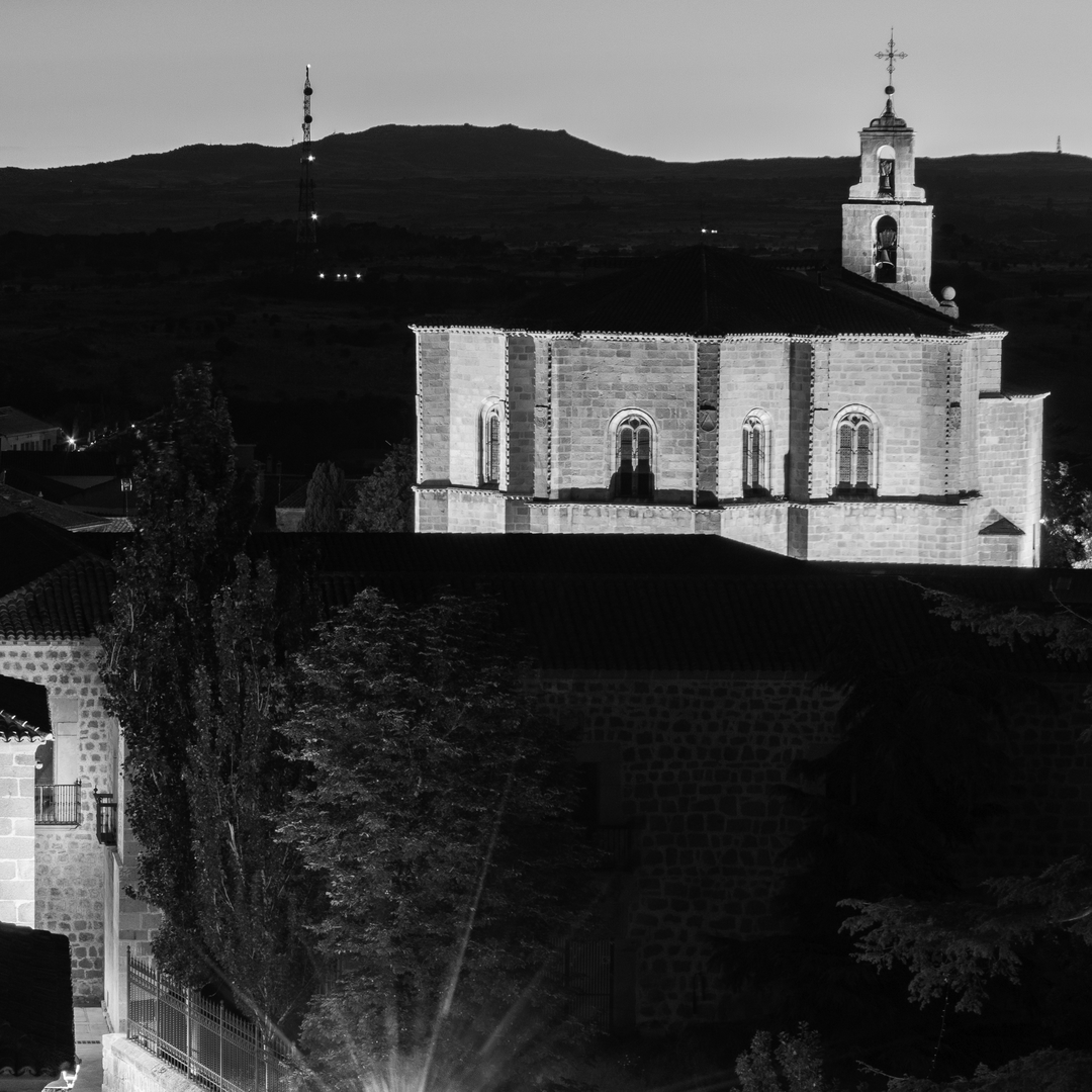  Serene majesty of the church in Mosen Rubí, Ávila, Spain. Artificial lighting reveals architectural details such as arched windows and a cross-topped bell tower, standing quietly against the softly lit hills and communication tower in the distance. Framed by trees and rooftops in the foreground, the image emphasizes the contrast between historical spirituality and modern presence under a dusky sky. (c) pmartinasi