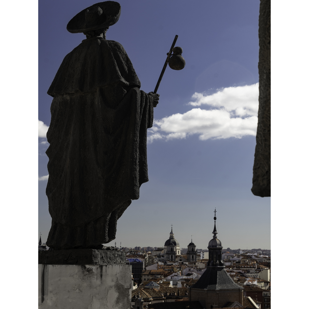  A bronze statue of Saint James (Santiago) stands atop the Cathedral of La Almudena in Madrid, Spain, facing the skyline and baroque domes of the historic city under a bright blue sky. (c) pmartinasi