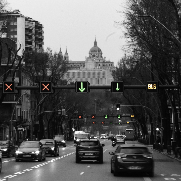 Street-level view of traffic moving through a wide avenue in Madrid, with illuminated traffic signals leading the eye towards the Almudena Cathedral in the distance, captured outdoors in a European city setting.