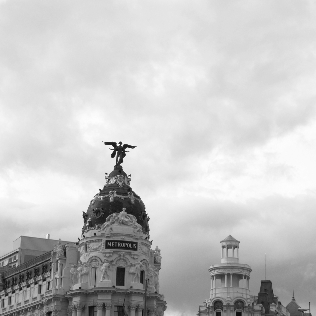  Black and white image of the iconic Metropolis Building on Gran Vía in Madrid, Spain, featuring ornate Beaux-Arts architecture with the famous winged statue against a moody sky backdrop. (c) pmartinasi