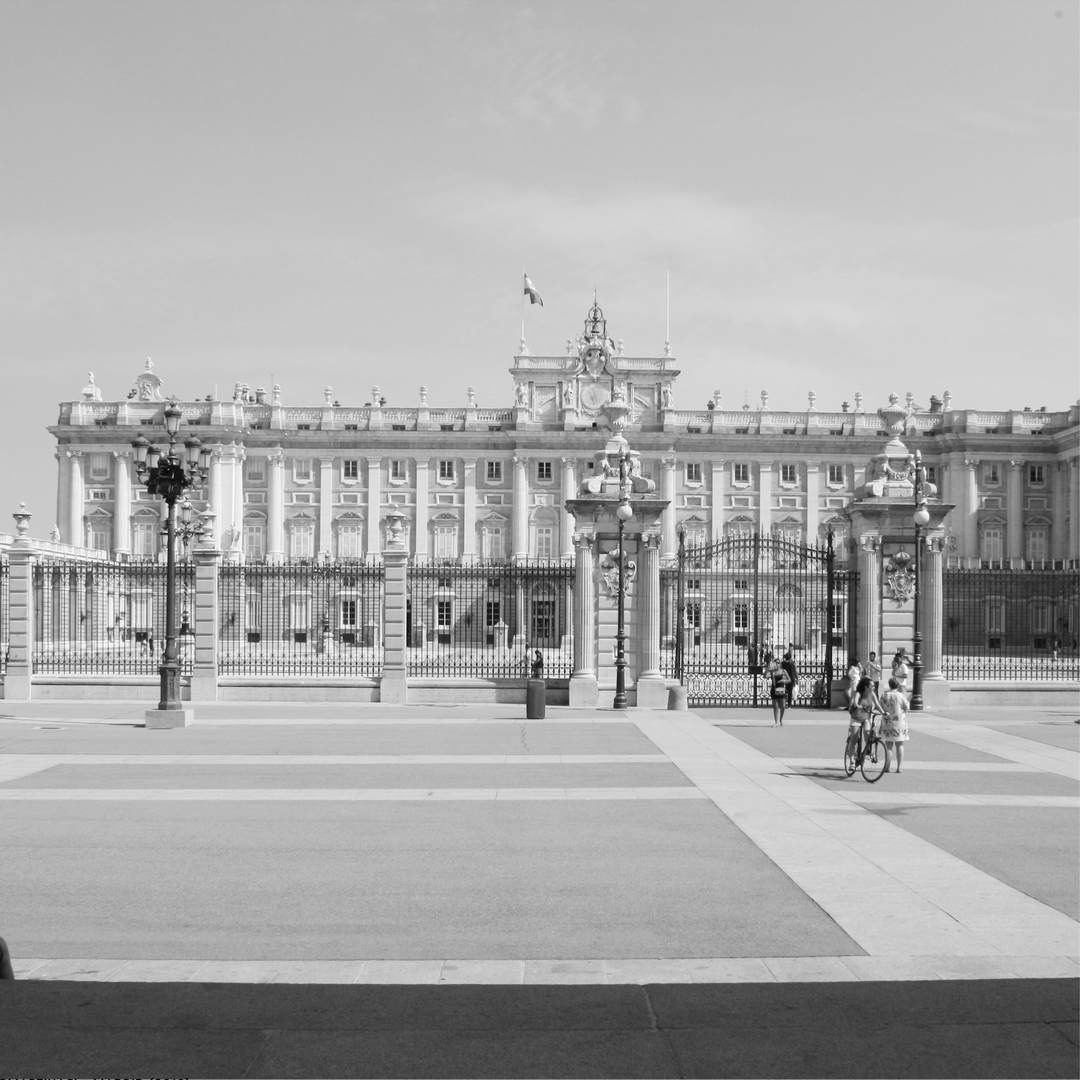 A timeless black and white photograph of the Royal Palace of Madrid (Palacio Real), capturing the grandeur of the architecture under a clear sky with pedestrians and cyclists animating the forecourt. (c) pmartinasi